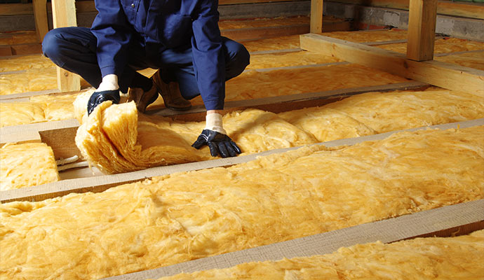 A technician in blue coveralls installing yellow fiberglass insulation in an attic