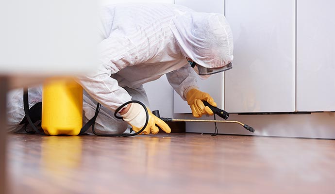 Professional in full protective gear disinfecting the floor area beneath a cabinet