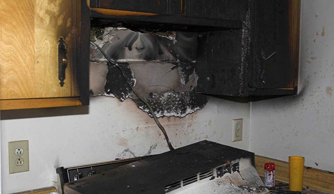 Fire damaged kitchen cabinets and wall with burned surfaces melted materials and heavy soot around stove area