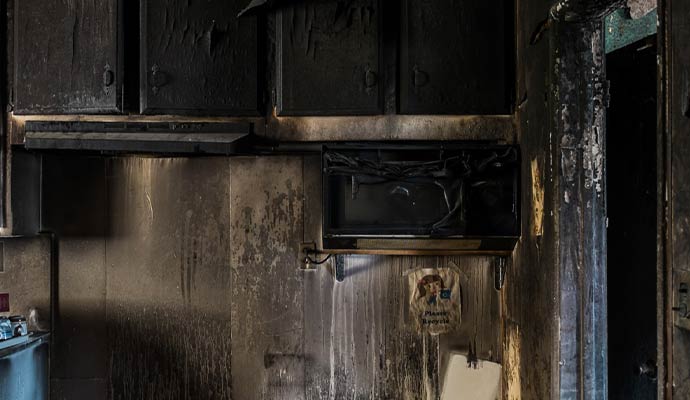 Severely fire damaged kitchen with charred cabinets smoke stains melted appliances and heavy soot buildup on walls