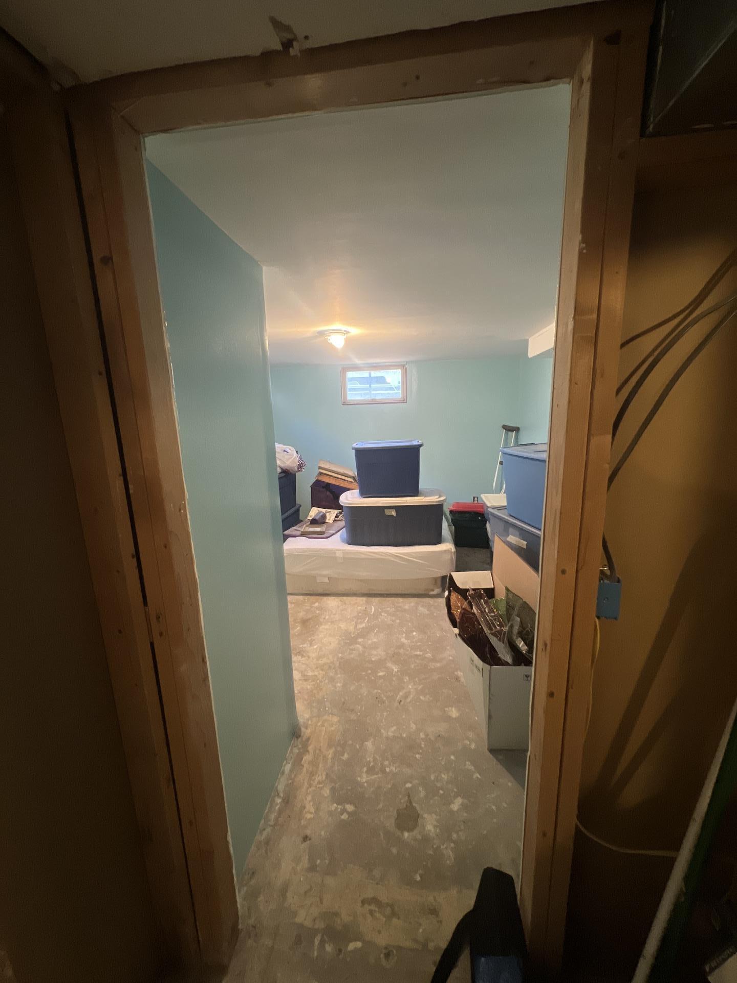 View into a basement bedroom from a doorway, showing a concrete floor affected by sewage contamination and various stored items.