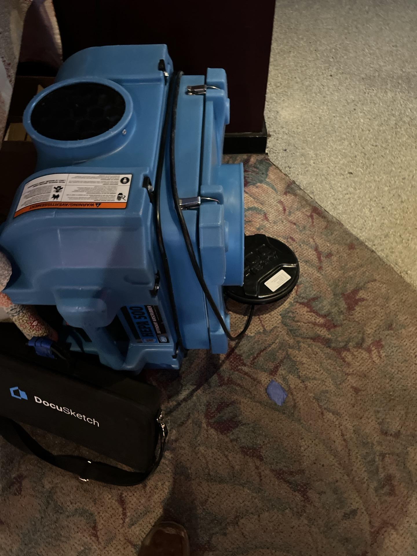 A blue air mover (blower fan) deployed on a patterned carpet for drying, with a DocuSketch kit visible nearby, indicating mitigation in progress.