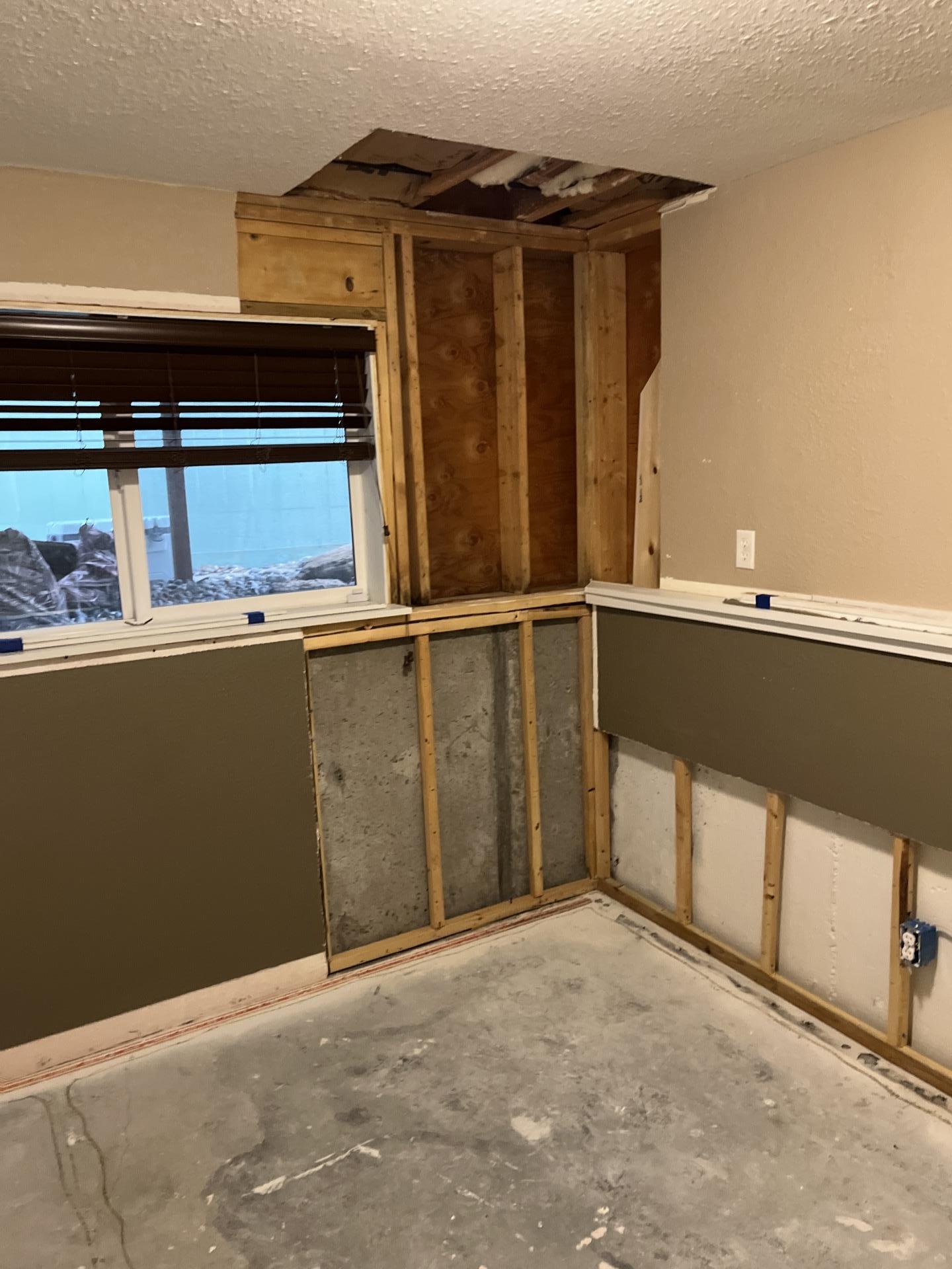 Interior room during water damage mitigation with drywall removed, exposing wooden wall studs, a concrete foundation, a window, and a section of the ceiling opened up.