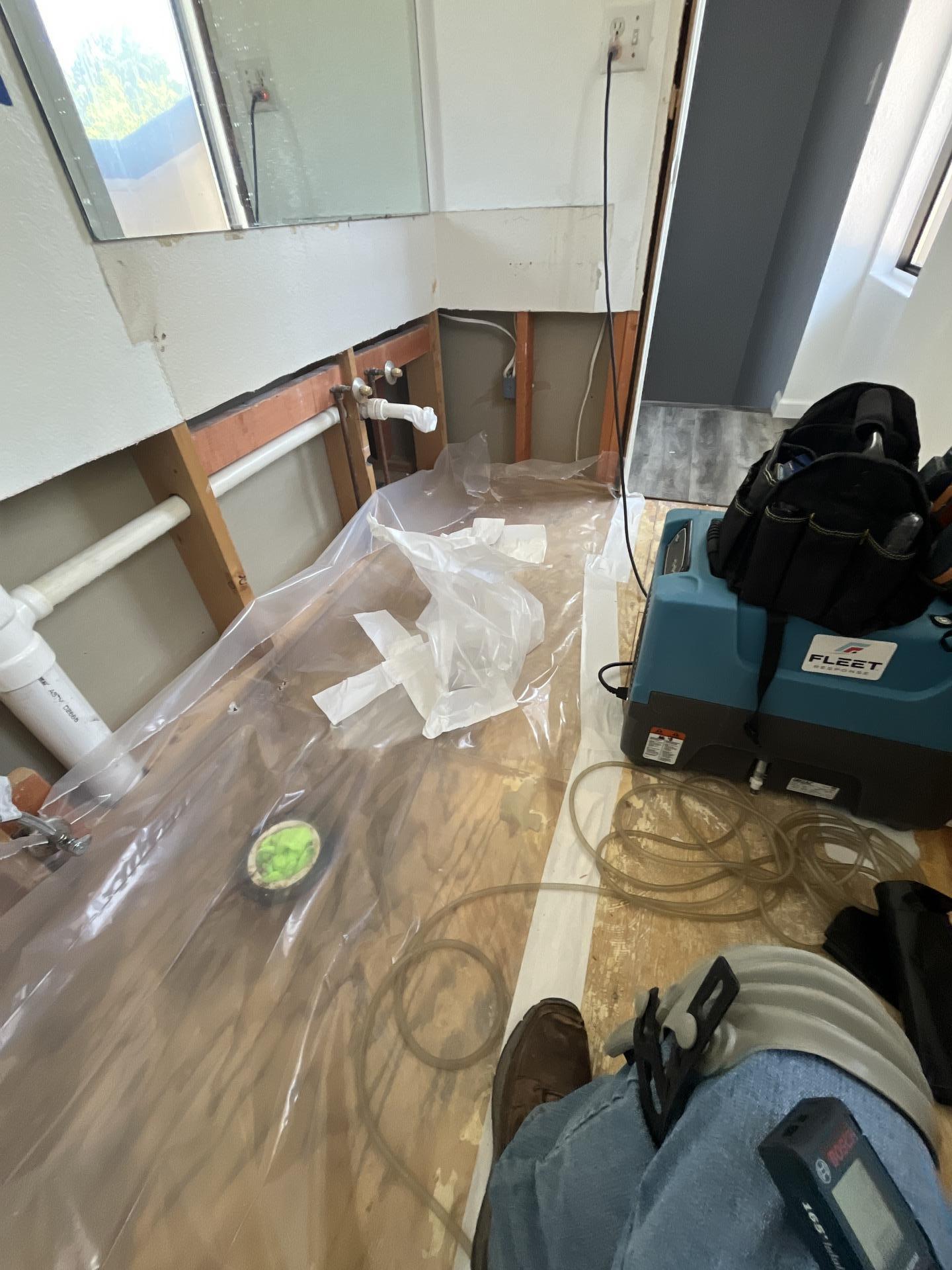 Bathroom water damage restoration in progress, featuring plastic sheeting, exposed framing, and professional drying equipment with a technician's tools.